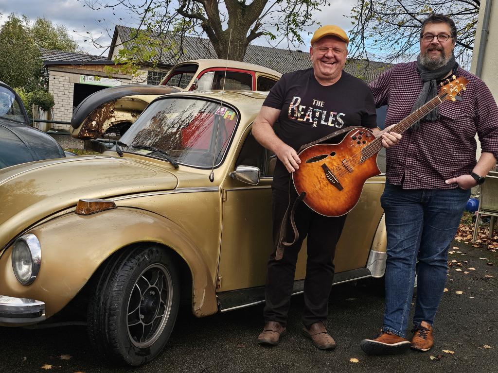 Thomas Küster und Michael Suermann stehen neben einem goldfarbenen VW Käfer. Einer der beiden hält eine Gitarre in der Hand, beide lächeln in die Kamera. Im Hintergrund sind weitere klassische Fahrzeu